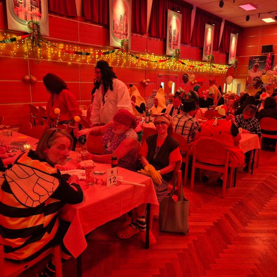 Ein Speisesaal mit rotem Thema ist mit Gästen gefüllt. Mehrere Personen sitzen an Tischen, einige tragen Kostüme. Ein Mann in Piratenkostüm steht neben einer Frau mit roter Bandana.