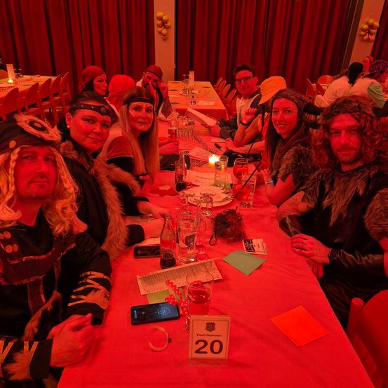 Eine Gruppe von Menschen in Kostümen sitzt an einem langen Tisch. Der Tisch ist rot gedeckt, mit verschiedenen Gegenständen wie Kerzen, Gläsern und einem Schild mit der Nummer 20. Die Umgebung scheint eine Party oder Zusammenkunft mit gedämpfter Beleuchtung zu sein.