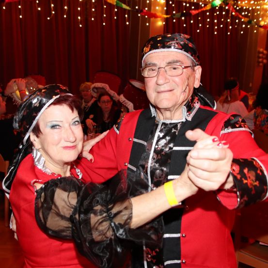 Ein älteres Paar, gekleidet in Rot und Schwarz, tanzt in einer festlichen Umgebung mit anderen um sie herum, unter dekorativen Lichtern.
