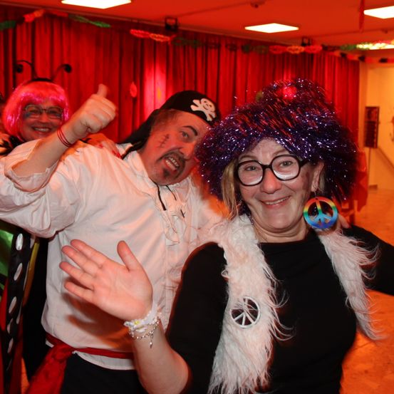 Drei Erwachsene in Kostümen auf einer Party. Ein Mann in einem Piratenhut gibt Daumen hoch, während eine Frau in einem Friedenszeichenhut lächelt. Eine weitere Person mit rosa Haaren steht hinter ihnen.