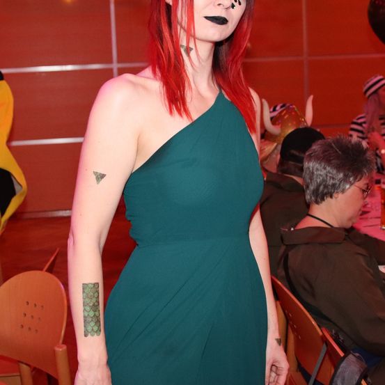 Eine Frau mit roten Haaren trägt ein türkisfarbenes Kleid und sichtbare Tattoos an den Armen steht in einem Raum mit anderen.