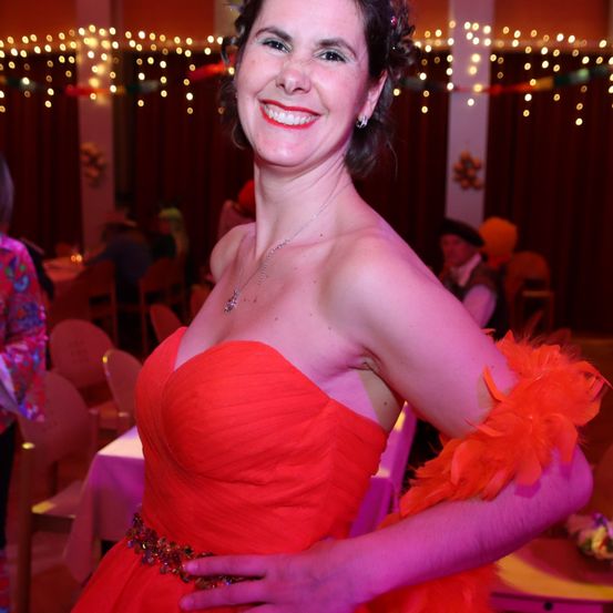 Eine Frau in einem roten Kleid und mit federigen Accessoires posiert für ein Foto bei einer festlichen Veranstaltung, mit Lichterketten und Menschen im Hintergrund.