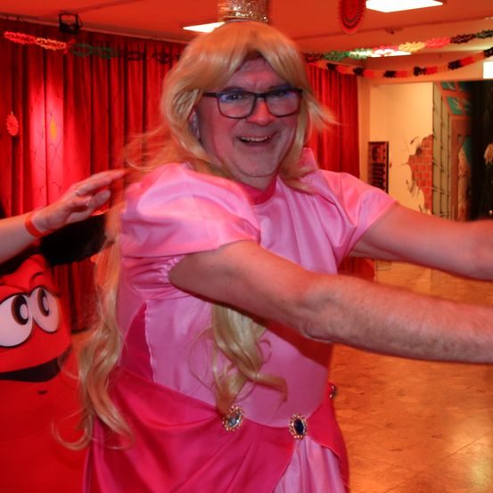 Eine Person in einem rosa Kleid und blonder Perücke mit Krone tanzt in einem Raum mit roten Vorhängen und Dekorationen.