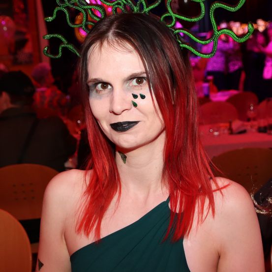 Eine Frau mit roten Haaren, grüner Kopfbedeckung und Gesichtsbemalung posiert in einem überfüllten Raum.