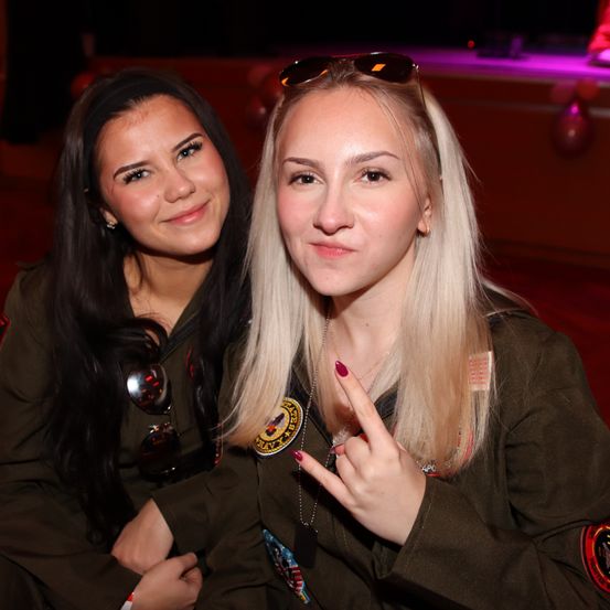 Zwei Frauen in Militäruniformen lächeln und posieren für ein Foto, wobei eine eine Friedensgeste macht. Sie stehen eng beieinander in einem schwach beleuchteten Raum.