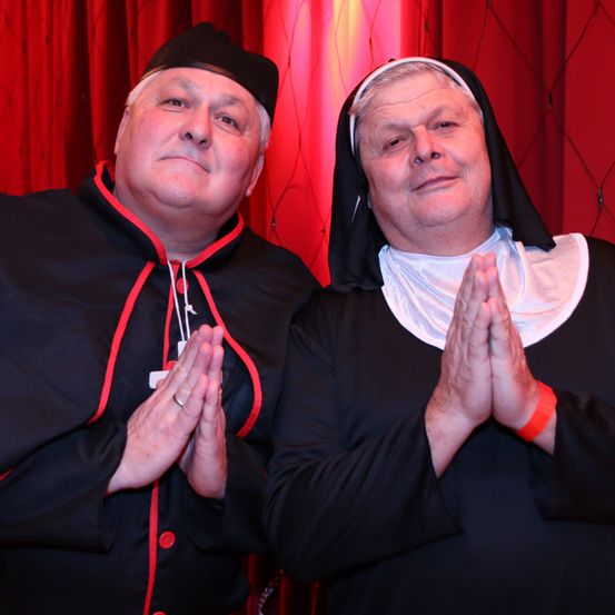 Zwei Männer in schwarzer religiöser Kleidung posieren für ein Foto vor einem roten Vorhang. Einer trägt einen schwarzen Umhang mit rotem Besatz und einen schwarzen Hut, der andere ein schwarzes Nonnengewand.