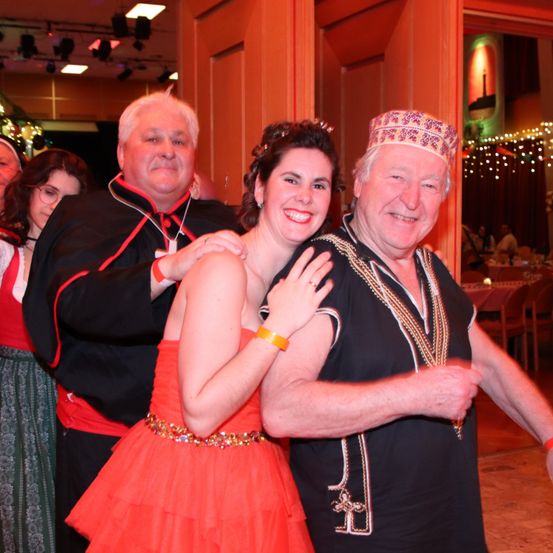 Eine Frau in einem orangefarbenen Kleid und ein Mann in einem schwarzen Kleid mit einer Halskette lächeln in die Kamera. Sie stehen neben einem Mann in einem schwarzen Gewand und einer Frau in einem grünen Kleid. Sie befinden sich auf einer Party mit einer Holztür und einer dekorierten Wand.