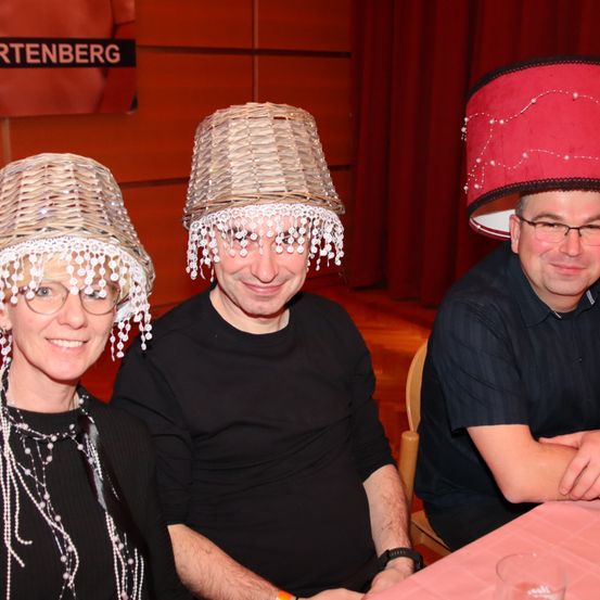 Drei Menschen mit ungewöhnlichen Hüten sitzen an einem Tisch. Die Frau trägt eine Perlenkette. Der Mann rechts trägt eine Brille. Hinter ihnen ist ein roter Vorhang.