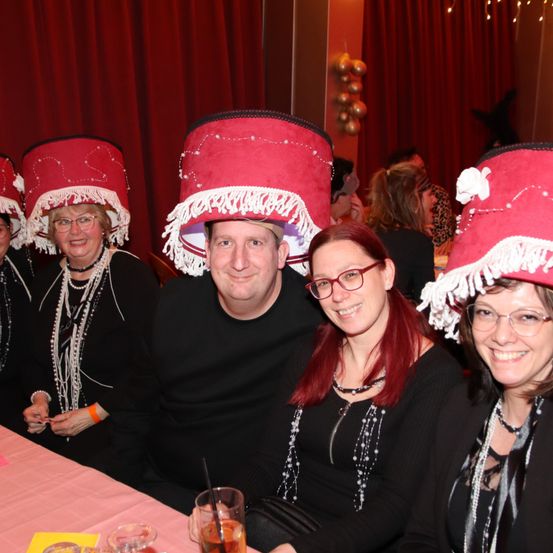 Eine Gruppe von Menschen posiert für ein Foto und trägt rote Hüte. Sie sitzen um einen Tisch herum, einer der Männer hält ein Getränk. Im Hintergrund befinden sich rote Vorhänge.