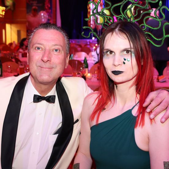 Ein Mann und eine Frau in formeller Kleidung posieren für ein Foto bei einer Veranstaltung. Der Mann trägt eine Fliege und die Frau hat rote Haare und grünes Gesichtsmake-up. Sie sind von Tischen und Stühlen umgeben.