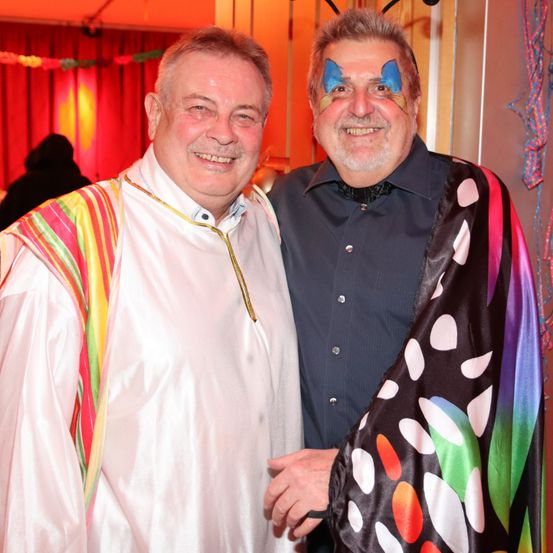 Zwei Männer lächeln für ein Foto. Einer trägt einen weißen Umhang und der andere hat bunte Schmetterlings-Gesichtsbemalung. Dahinter ist ein roter Vorhang.