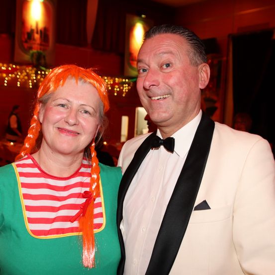 Ein lächelnder Mann in einem weißen Frack posiert neben einer Frau in einem rot-weiß gestreiften Hemd und einer orangefarbenen Perücke. Sie sind bei einer Veranstaltung im Innenbereich mit festlichen Lichtern im Hintergrund.
