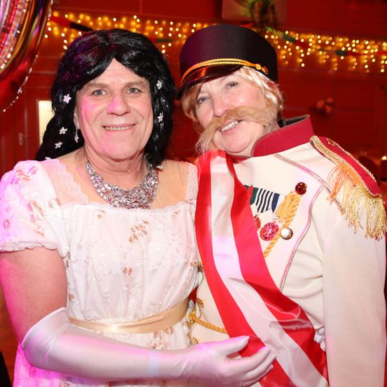 Zwei Menschen in Kostümen lächeln und posieren für ein Foto. Eine Frau in einem rosa Kleid mit Blumenmuster und ein Mann in einem weißen Kostüm mit einem roten Schal.