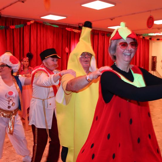 Eine Gruppe von Erwachsenen in Kostümen tanzt zusammen. Einer trägt eine Bananen-Verkleidung, ein anderer ein Erdbeerkostüm und ein Dritter einen Pilzhut. Sie befinden sich in einem Raum mit roten Vorhängen und festlichen Dekorationen.