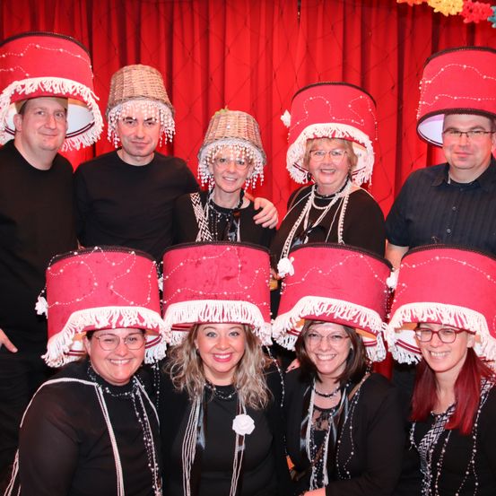 Eine Gruppe von Menschen, die rote Hüte mit dekorativen Quasten und Perlen tragen, posiert für ein Foto vor einem roten Vorhang. Einige tragen Brillen und Halsketten. Zwei Männer, einer mit Brille, stehen an den Seiten.