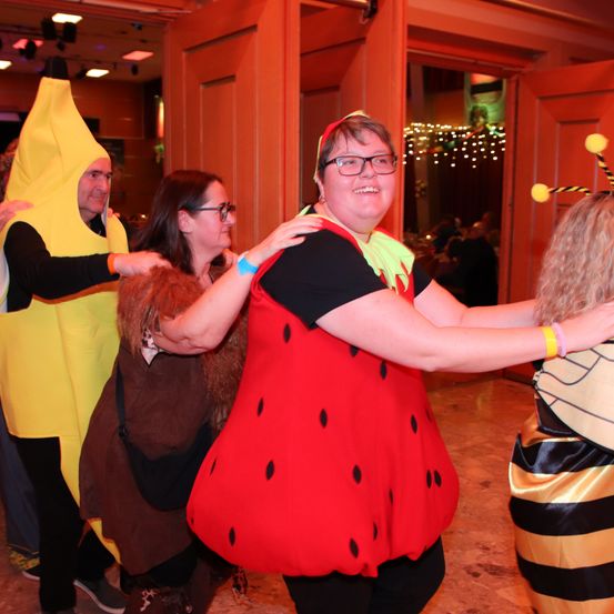 Mehrere Erwachsene in Kostümen tanzen in einem Raum mit warmer Beleuchtung. Eine Person trägt ein Wassermelonenkostüm, während andere in Bananen- und Bienenkostümen sind.
