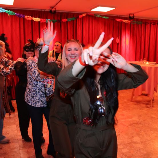 Zwei Frauen in Militäruniformen tanzen auf einer Party, eine macht ein Friedenszeichen mit ihren Händen. Dahinter tanzen Menschen, und ein Tisch mit Tischdecke ist aufgebaut.