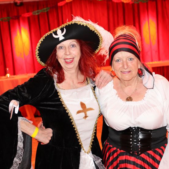 Zwei Frauen in Piratenkostümen, eine mit einem Hut mit Feder und einem Kreuz auf der Brust, stehen zusammen vor einem roten Vorhang.