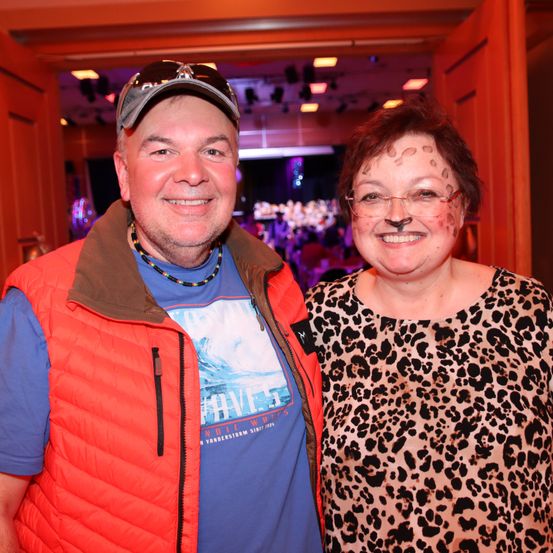 Ein Mann und eine Frau stehen gemeinsam und lächeln für ein Foto. Der Mann trägt eine rote Weste und eine Kappe. Die Frau hat Leoparden-Make-up im Gesicht. Sie scheinen sich bei einer Veranstaltung zu befinden.