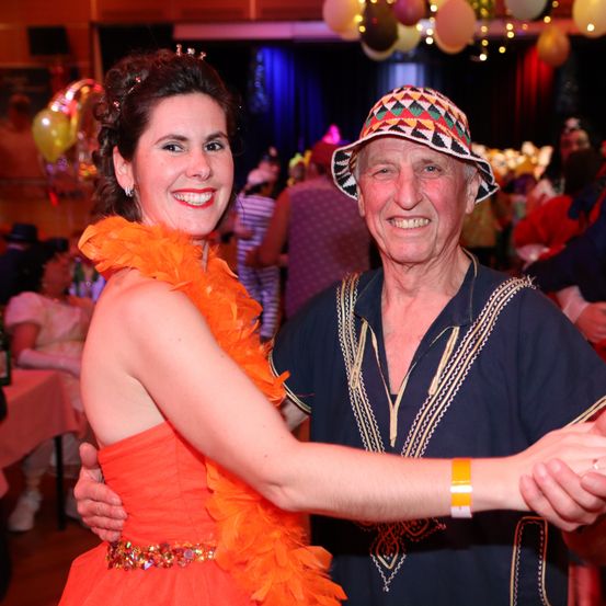 Ein lächelndes Paar tanzt bei einer festlichen Veranstaltung zusammen, die Frau trägt ein orangefarbenes Federkleid und der Mann einen bunten Hut und ein blaues Hemd.
