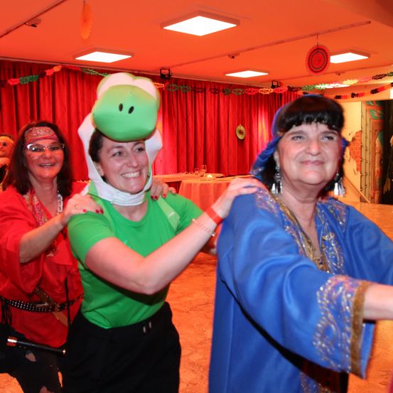 Drei Frauen tanzen in einem dekorierten Raum. Eine trägt einen grünen Hut, eine andere ein rotes Kleid und die dritte ein blaues Gewand. Hinter ihnen steht ein Tisch mit einer weißen Tischdecke und Weingläsern.