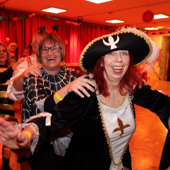 Mehrere Erwachsene in festlicher Kleidung, darunter eine Frau in einer Piraten-Kostüm, tanzen zusammen auf einer Tanzfläche mit roten Vorhängen und festlichen Dekorationen.