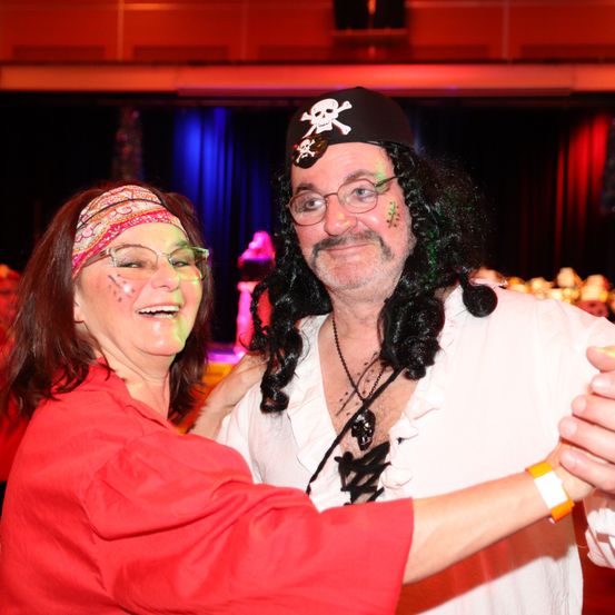 Eine lächelnde Frau in einem roten Shirt und ein Mann in einer Piraten-Verkleidung tanzen gemeinsam in einer festlichen Umgebung.