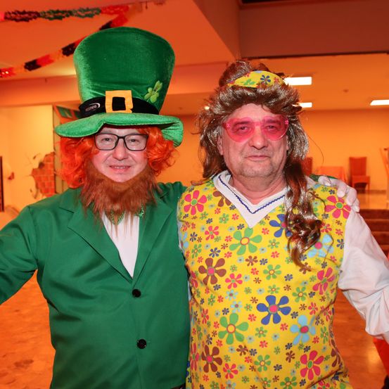 Zwei Männer sind in Kostümen gekleidet. Einer trägt einen grünen St. Patrick's Day Hut, während der andere ein gelbes Blumenhemd trägt. Sie haben beide Perücken und lächeln.