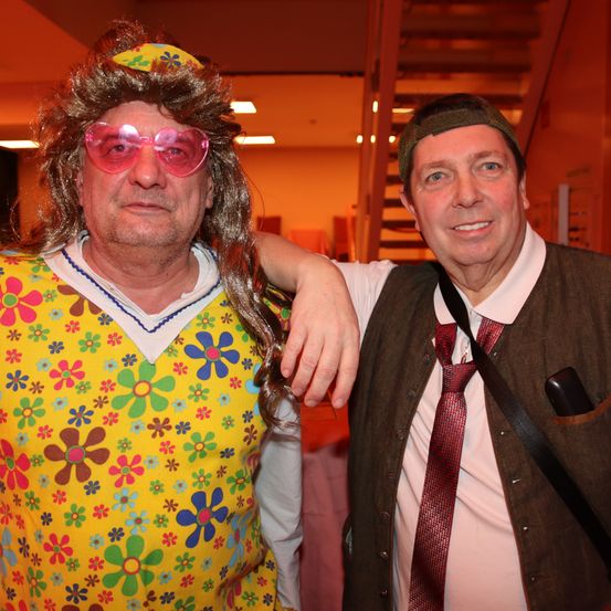 Zwei Männer posieren für ein Foto, einer in einem farbenfrohen Blumenhemd und Sonnenbrille, der andere in einer Weste und Krawatte mit einer Tasche an der Schulter.