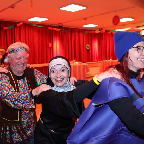 Drei Personen in einem Raum mit roten Vorhängen, ein Mann in bunter Kleidung, eine Frau in einer Nonnentracht und eine weitere Frau in einem blauen Kostüm, alle lächeln und tanzen gemeinsam.