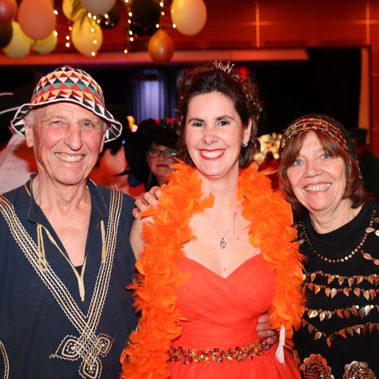 Drei Menschen lächeln in die Kamera. Die Frau in der Mitte trägt eine Krone und eine orangefarbene Federboa. Der Mann links trägt einen Hut. Die Frau rechts trägt ein schwarzes Shirt.