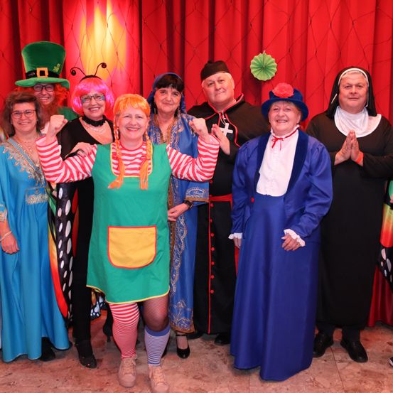 Eine Gruppe von Menschen in farbenfrohen Kostümen posiert für ein Foto vor einem roten Vorhang. Einige tragen Hüte, Brillen und Halsketten. Eine Person in einem grünen Kleid hat die Hände erhoben.