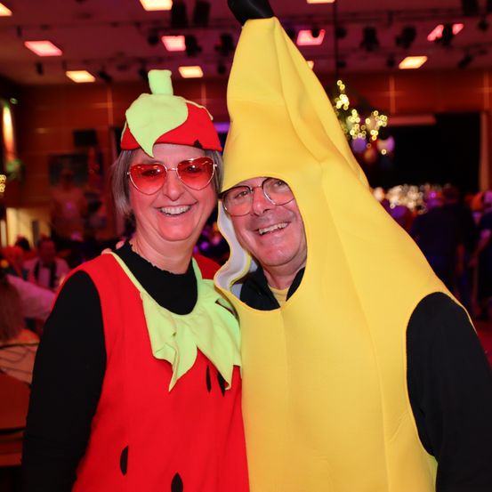 Ein Mann und eine Frau in Obstkostümen lächeln in einem überfüllten Raum in die Kamera.