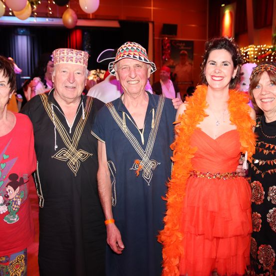 Fünf Menschen sind auf einer Party und lächeln für ein Foto. Zwei Männer tragen traditionelle Kleidung und Hüte. Eine Frau in einem orangefarbenen Kleid trägt einen Federboa. Der Hintergrund ist mit Ballons und Lichtern dekoriert.