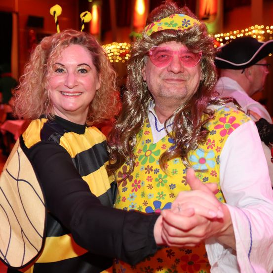 Eine Frau in einem Bienenkostüm und ein Mann in einem Flower-Power-Hemd lächeln und tanzen bei einer Party zusammen.