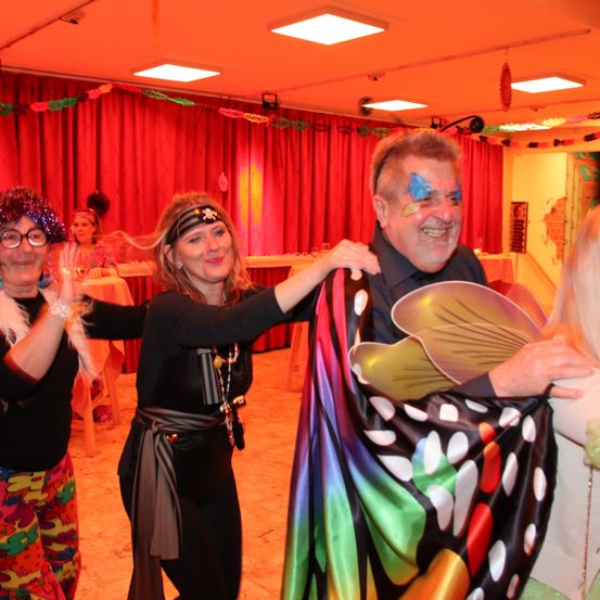 Eine Gruppe von Menschen in Kostümen tanzt in einem Raum mit einem roten Vorhang. Die Frau auf der linken Seite trägt eine Brille. Der Mann in der Mitte trägt ein Schmetterlingskostüm. Die Frau auf der rechten Seite trägt ein grünes Kleid.
