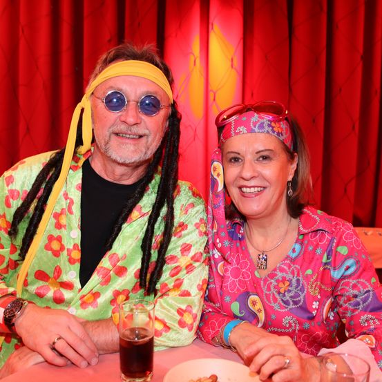 Ein Mann und eine Frau in farbenfrohen, blumigen Kleidern sitzen an einem Tisch, lächeln, mit Getränken und Essen vor ihnen und einem roten Vorhang dahinter.