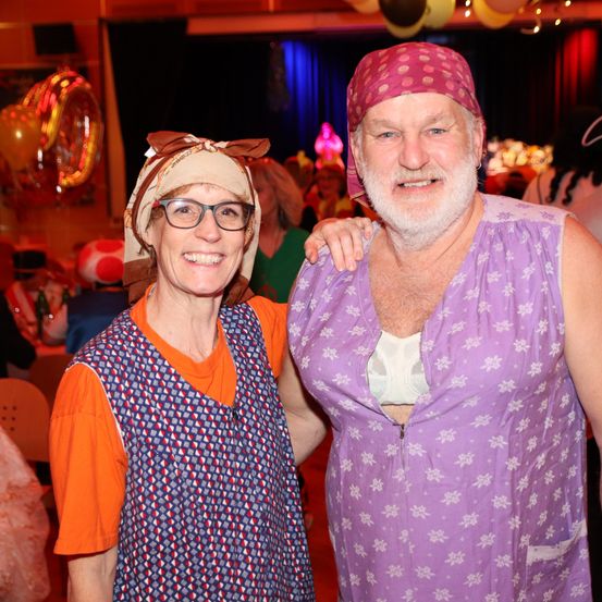 Ein älterer Mann und eine ältere Frau stehen eng beieinander und lächeln in die Kamera. Sie tragen passende Kostüme auf einer Party, mit Luftballons und Lichtern im Hintergrund.