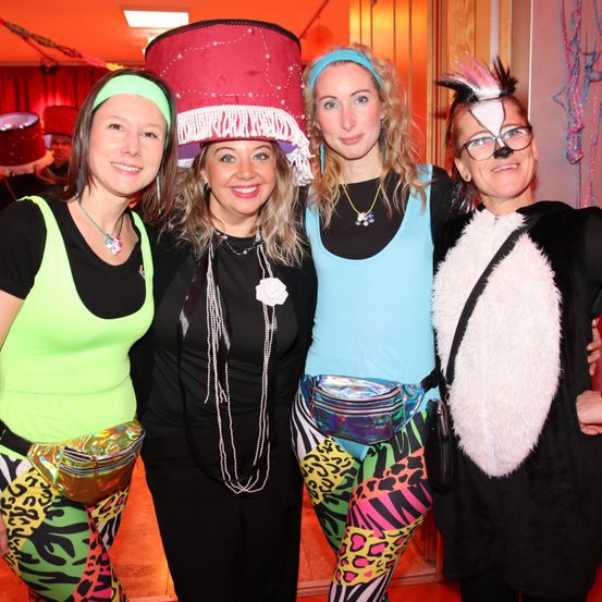 Vier Frauen in farbenfrohen und einzigartigen Outfits posieren bei einer Party für ein Foto, eine trägt einen großen Hut.
