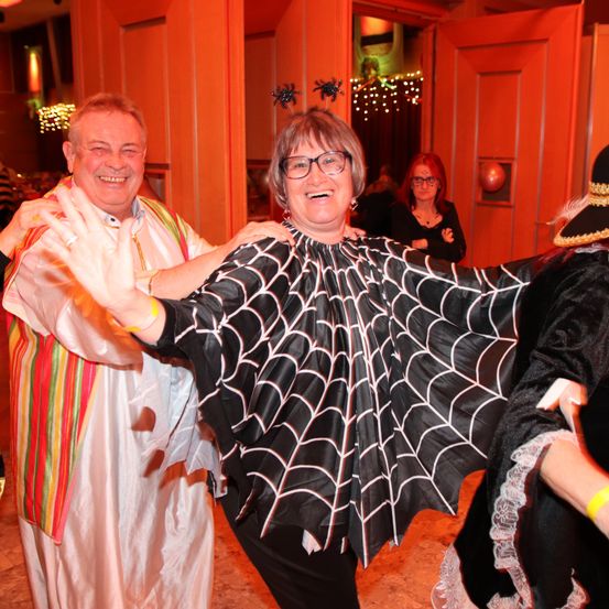 Eine Frau in einem spinnenartigen Kostüm winkt in die Kamera, während ein Mann neben ihr ein farbenfrohes Gewand trägt. Dahinter steht eine weitere Frau mit verschränkten Armen. Der Raum ist mit Halloween-Elementen dekoriert.