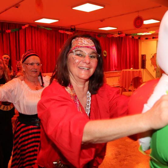 Eine Frau in einem roten Kleid und Stirnband tanzt mit einer Puppe. Hinter ihr lächelt eine Frau in einem gestreiften Kleid und Bandana. Sie befinden sich in einem Raum mit roten Vorhängen und einem Tisch mit Tischdecke.