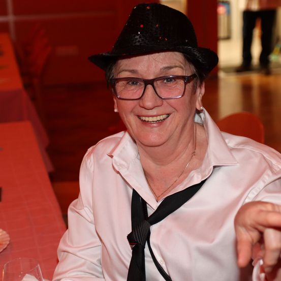 Eine Frau in einem weißen Hemd und schwarzem Hut lächelt zur Kamera bei einer Veranstaltung. Hinter ihr ist ein rot gestalteter Raum mit Tisch und Stühlen.