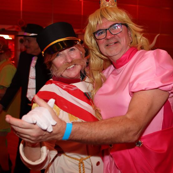 Zwei Erwachsene in Kostümen auf einer Party. Der Mann trägt ein rosa Kleid und eine Krone, während die Frau ein weißes und rotes Kostüm und einen schwarzen Zylinderhut trägt.