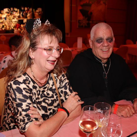 Ein älteres Paar sitzt zusammen an einem Tisch, die Frau mit Leopardenmusterkleidung und einer Katzen-Ohr-Haarband, lächelnd. Der Mann trägt ein schwarzes Outfit und hat Gesichtsbemalung. Sie sind umgeben von Gläsern und einer rosa Tischdecke.