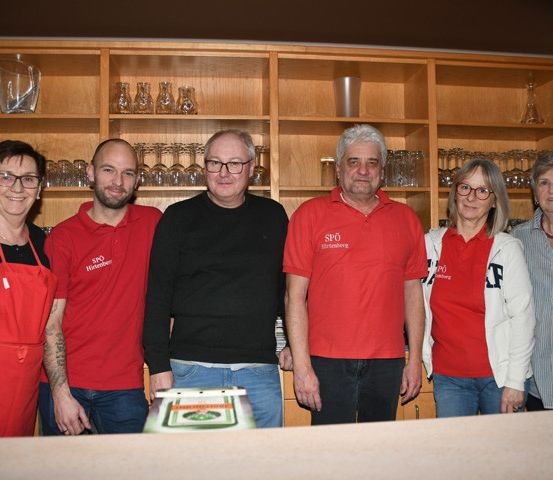 Eine Gruppe von Menschen, die rote Hemden und Schürzen tragen, posiert für ein Foto in einem Raum mit Holzregalen voller Gläser und einem Tisch mit einem Kuchen davor.