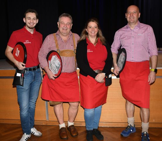 Vier Personen, die in Rot gekleidet sind, stehen zusammen, möglicherweise auf einer Bühne. Jede einzelne hält ein rotes Paddel und trägt ein rotes Schürzen. Hinter ihnen ist eine Holzkulisse und ein schwarzer Vorhang.