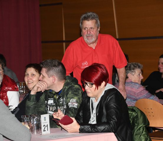 Eine Gruppe von Menschen sitzt an einem Esstisch, einige unterhalten sich, während ein Mann in einem roten Shirt hinter ihnen steht.