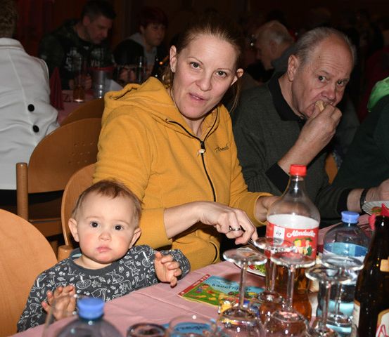 Eine Familie sitzt an einem Tisch, mit einem Baby davor, einer Frau in einer gelben Jacke und zwei älteren Personen dahinter. Auf dem Tisch stehen verschiedene Flaschen, Becher und Weingläser.