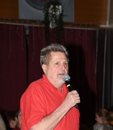 Ein älterer Mann in einem roten Hemd spricht in ein Mikrofon vor einer Menschenmenge.