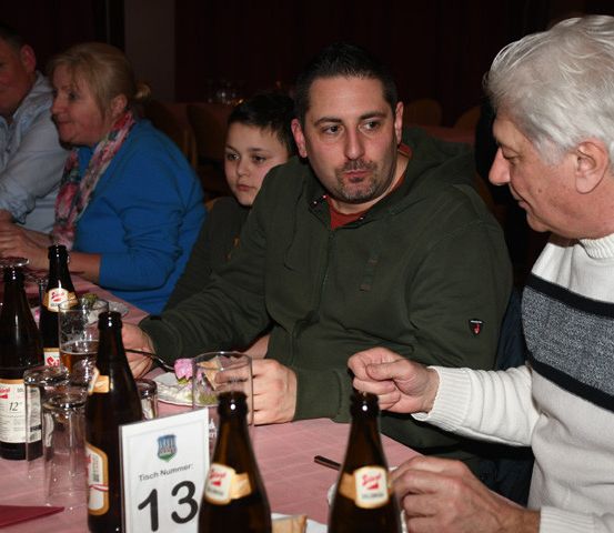 Mehrere Erwachsene und ein Kind sitzen an einem Tisch und genießen eine Mahlzeit mit verschiedenen Getränken und Gerichten. Der Mann im grünen Jackett führt ein Gespräch mit einem älteren Mann.